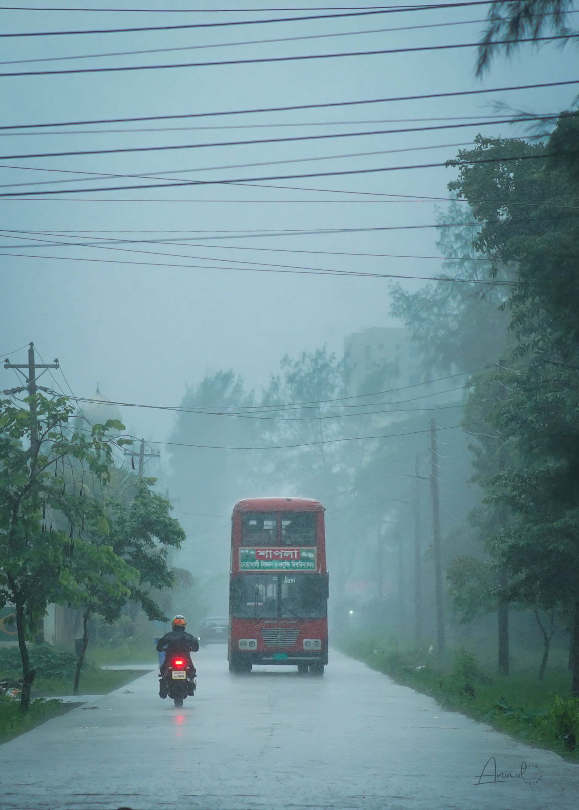 A serene street scene with a double-decker bus and motorbike on a misty monsoon day in Noakhali.