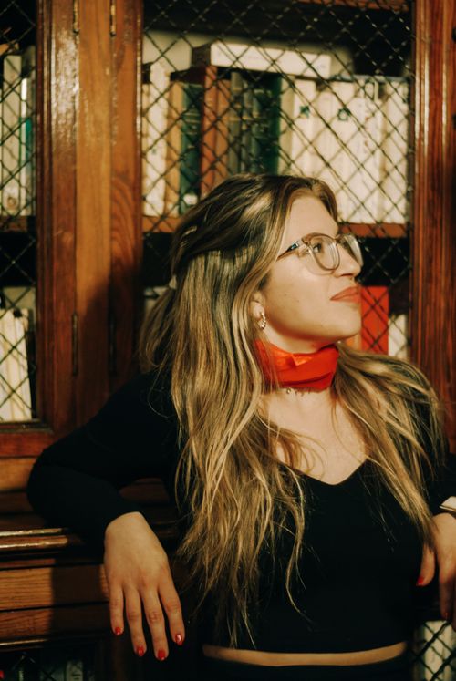 Free Stylish woman wearing glasses and red scarf, leaning on library bookshelf. Stock Photo
