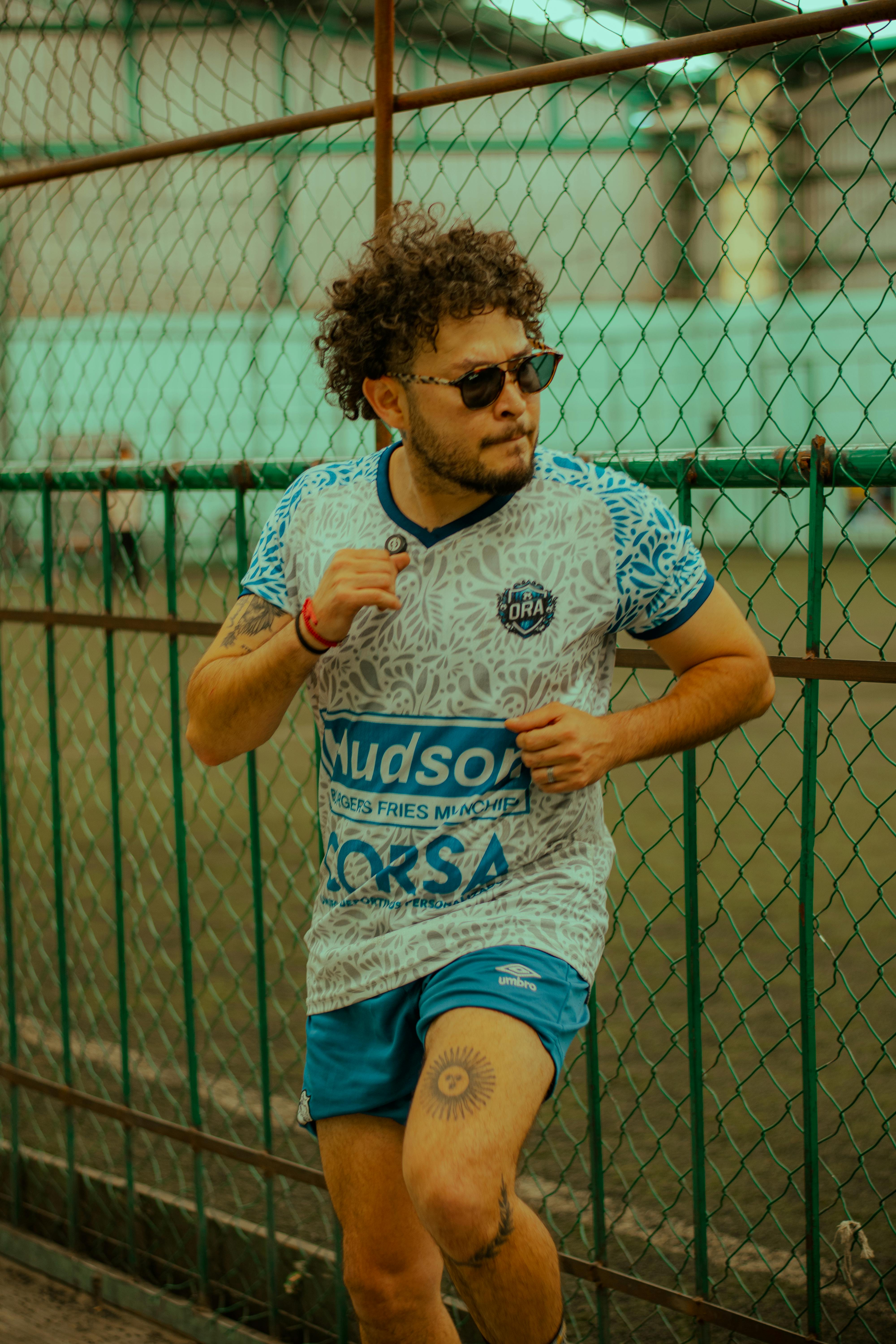 Energetic young male athlete running outside along a chain-link fence with a focused expression.