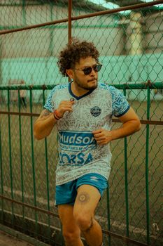 Energetic young male athlete running outside along a chain-link fence with a focused expression.