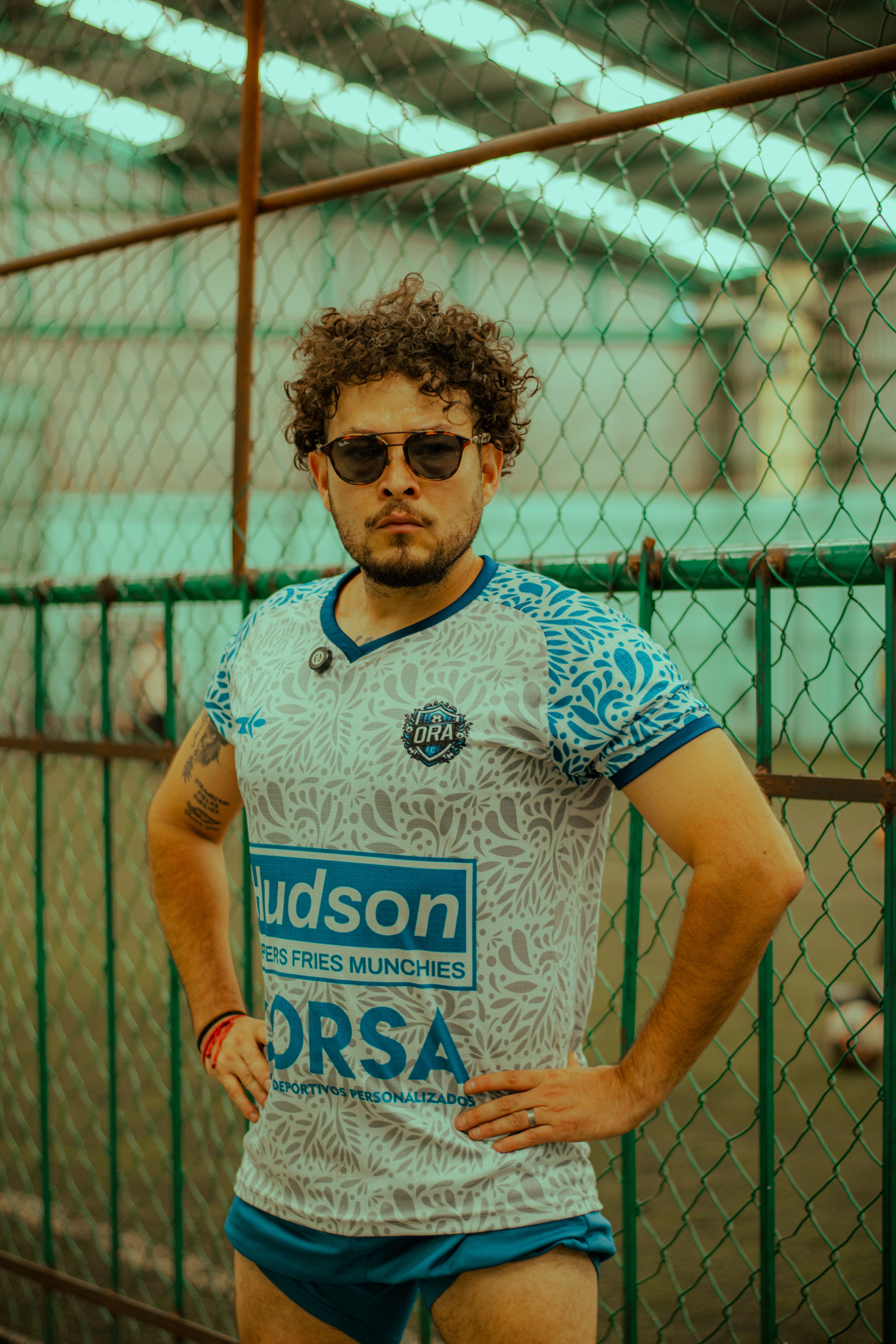 Confident man wearing printed jersey poses by a chain-link fence outdoors.