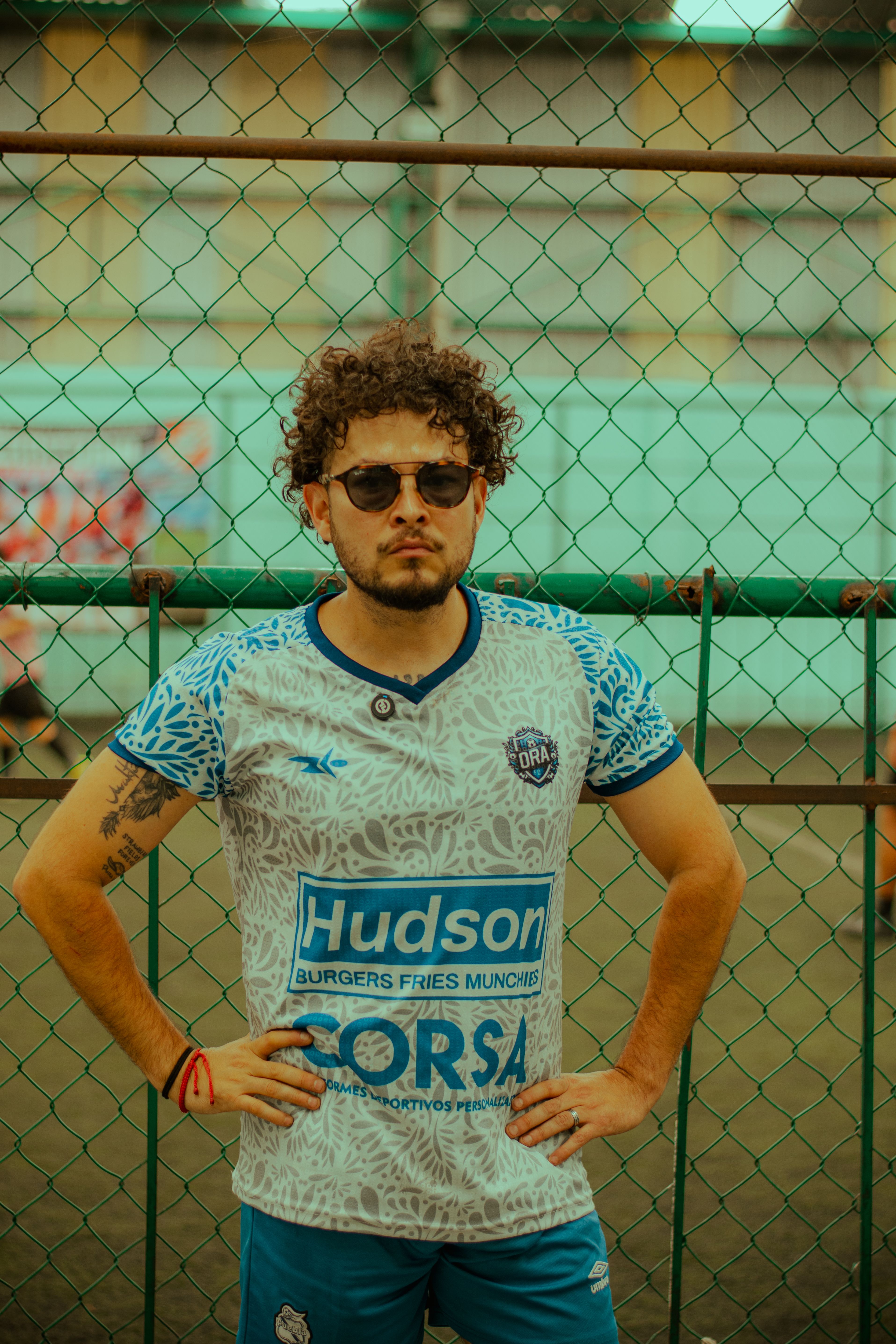 Confident male athlete wearing sunglasses and a sports jersey poses by a fence on a sunny day.