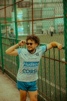 Casual urban lifestyle image of a young man in sportswear lounging against a fence.