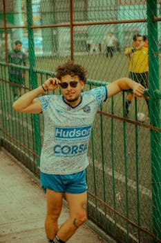A young man with sunglasses poses by a soccer field fence, embodying a sporty casual vibe.