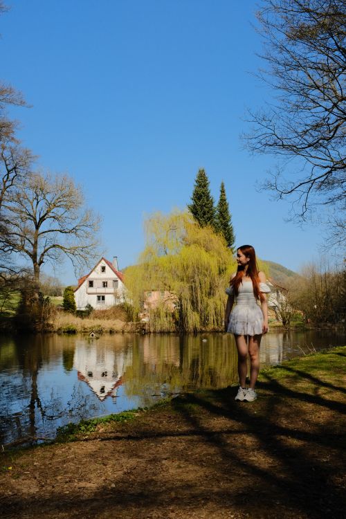 Free A woman stands by a serene lake with clear reflections and spring scenery. Stock Photo