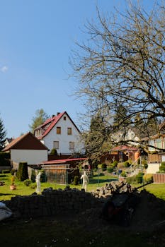 A peaceful garden with statues, trees, and houses under a clear blue sky.