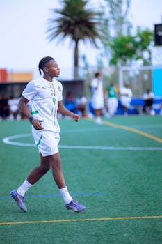 A teenage soccer player in uniform walking on a green field during a sunny day.