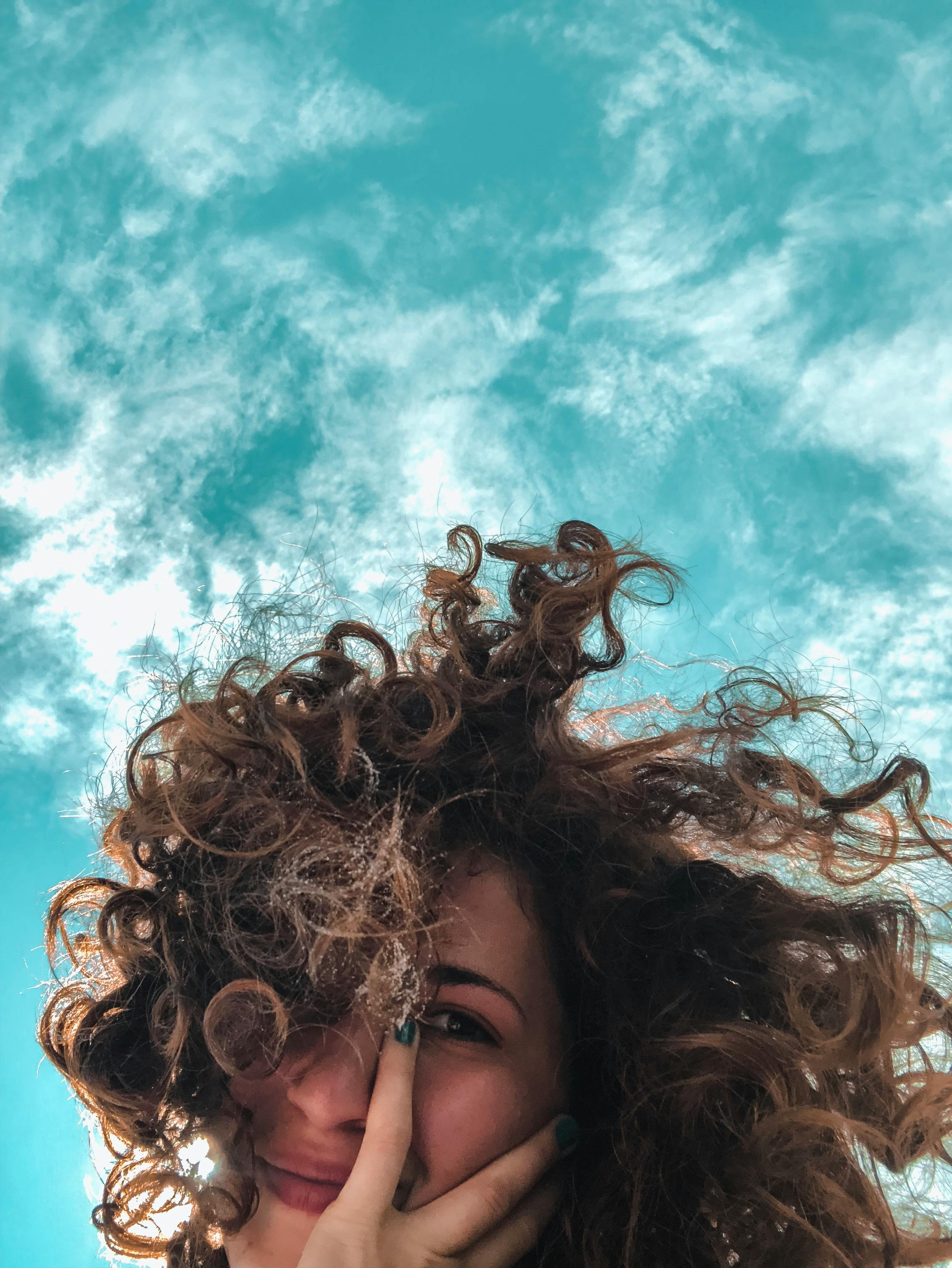 From below of charming young female with curly hair looking at camera with cloudy blue sky on background