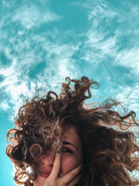 From below of charming young female with curly hair looking at camera with cloudy blue sky on background