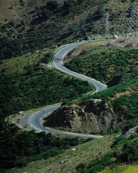 A winding road cuts through a lush green mountain landscape under a clear sky, offering a picturesque view.