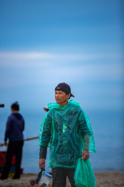 Free A man on the beach wearing a green rain poncho, holding gear, with a calm sea backdrop. Stock Photo