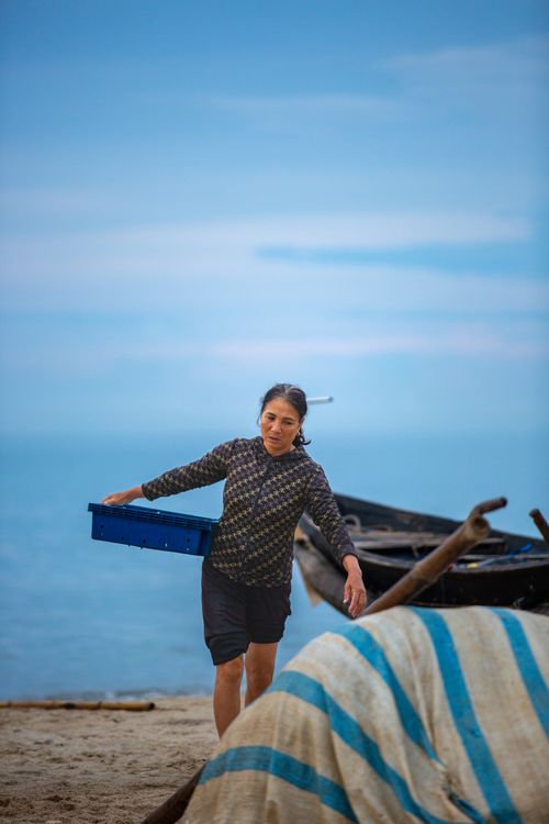 Free An Asian woman carries a basket near a boat on a serene beach, evoking a traditional lifestyle. Stock Photo