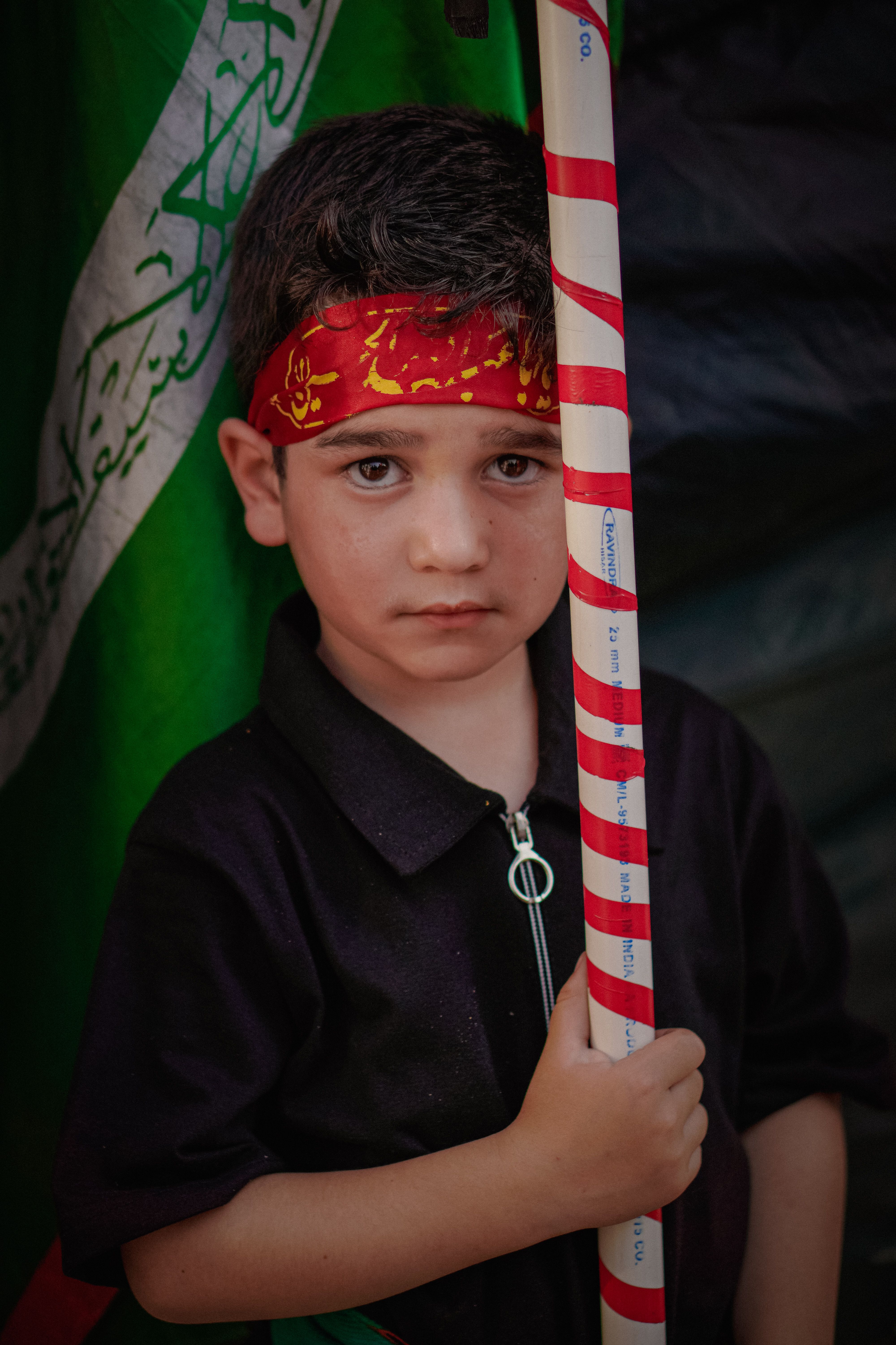 A child in a red headband holds a striped flag pole against a background featuring Arabic script.