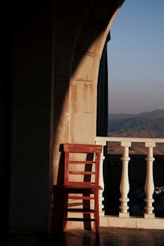 A wooden chair on a sunlit balcony overlooking a scenic mountain view at sunset.