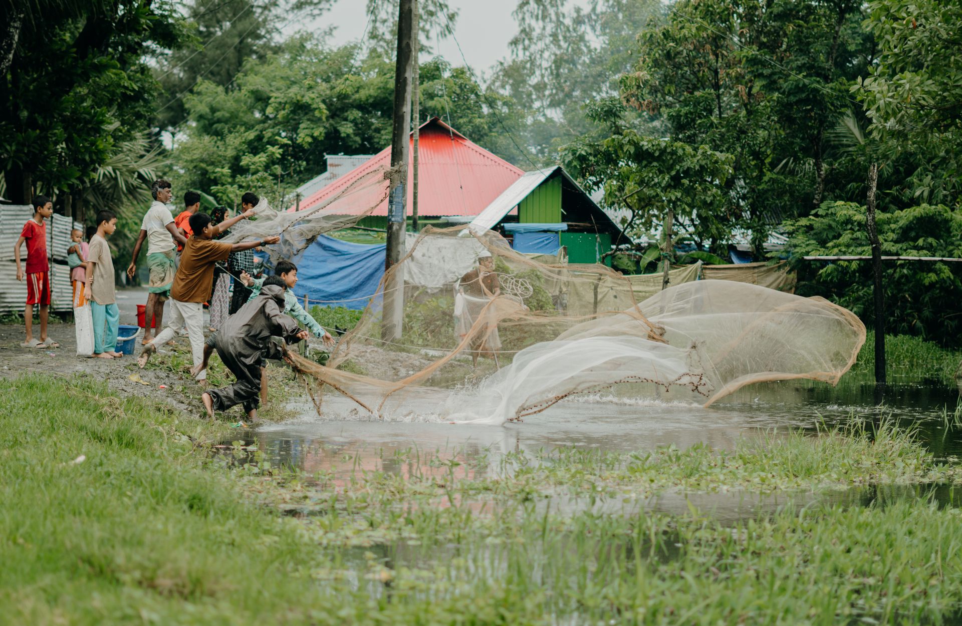 People casting fishing nets in a rural waterside village with lush greenery and colorful houses.