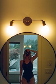 A woman in a swimsuit taking a mirror selfie indoors, capturing a scenic view outside.