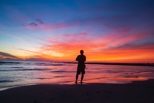 Free Silhouette of a person standing on a beach during a colorful sunset, reflecting over the ocean. Stock Photo