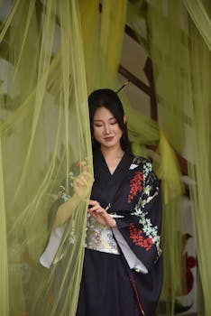 A serene portrait of a woman in a traditional kimono standing gracefully behind sheer yellow curtains.