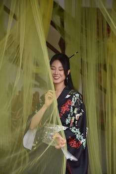 Elegant woman in a kimono gracefully standing behind delicate yellow curtains, exuding serene beauty.