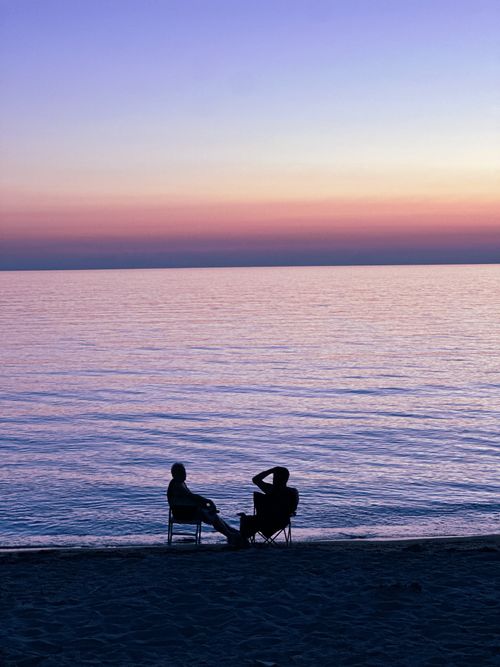 Free Silhouettes of two people seated at the beach during a serene sunset over calm ocean waters. Stock Photo