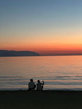 Two people sit on the beach at sunset, capturing the tranquil sea view with a smartphone.