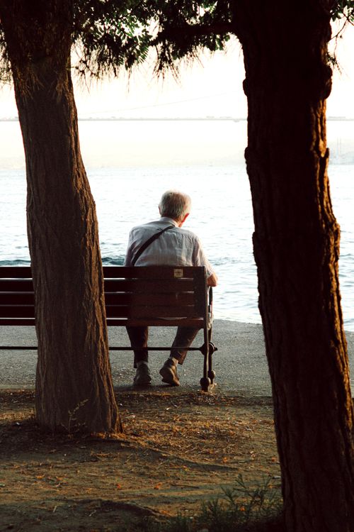 Free An elderly man sits on a bench by the waterfront, enjoying the sunset. Stock Photo