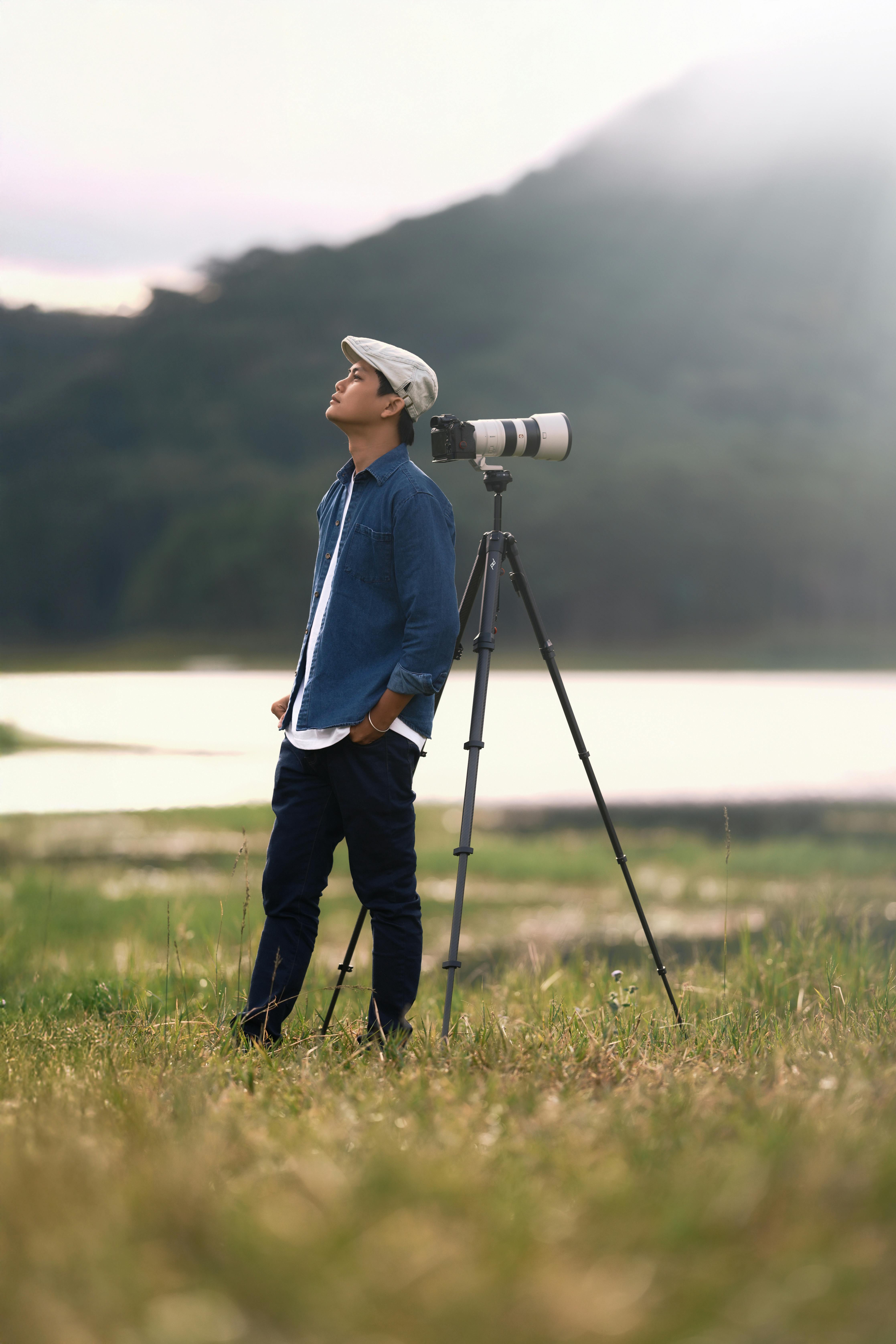 A photographer in the field observing nature beside a tripod-mounted camera with a scenic backdrop.
