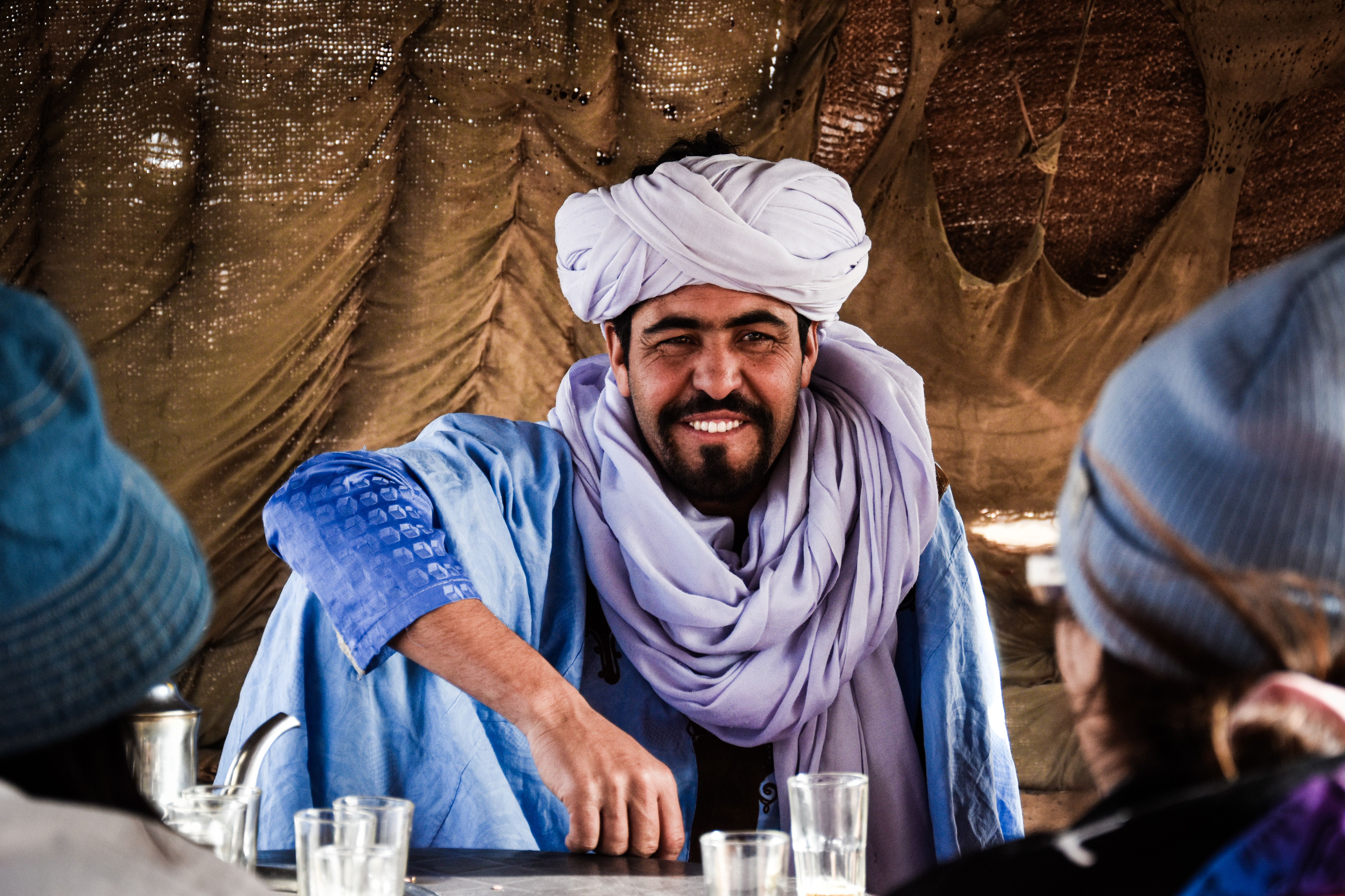 A man in a blue robe and white turban smiles inside a tent, offering a warm welcome.