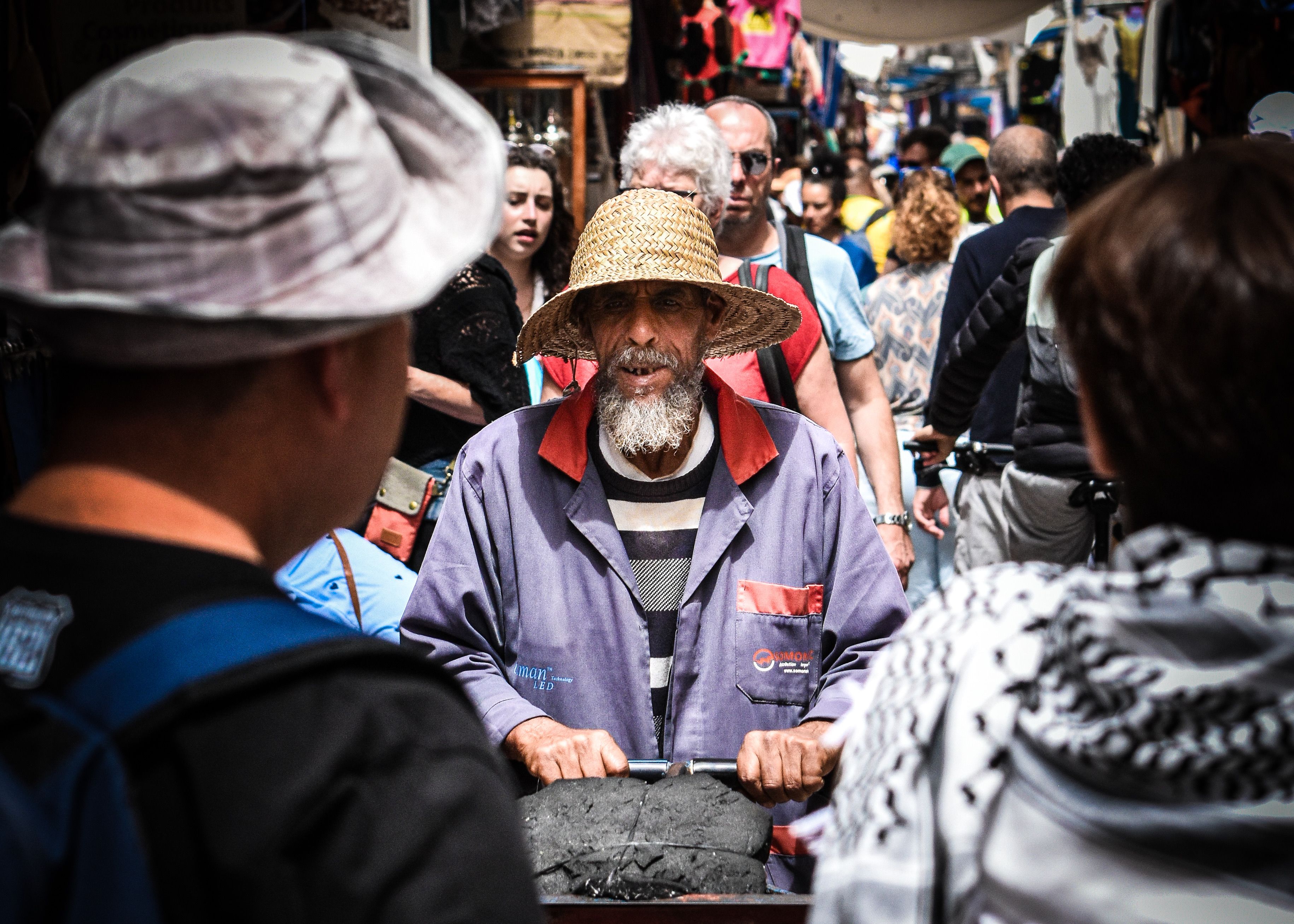 Elderly vendor with straw hat and cart in crowded outdoor market setting.