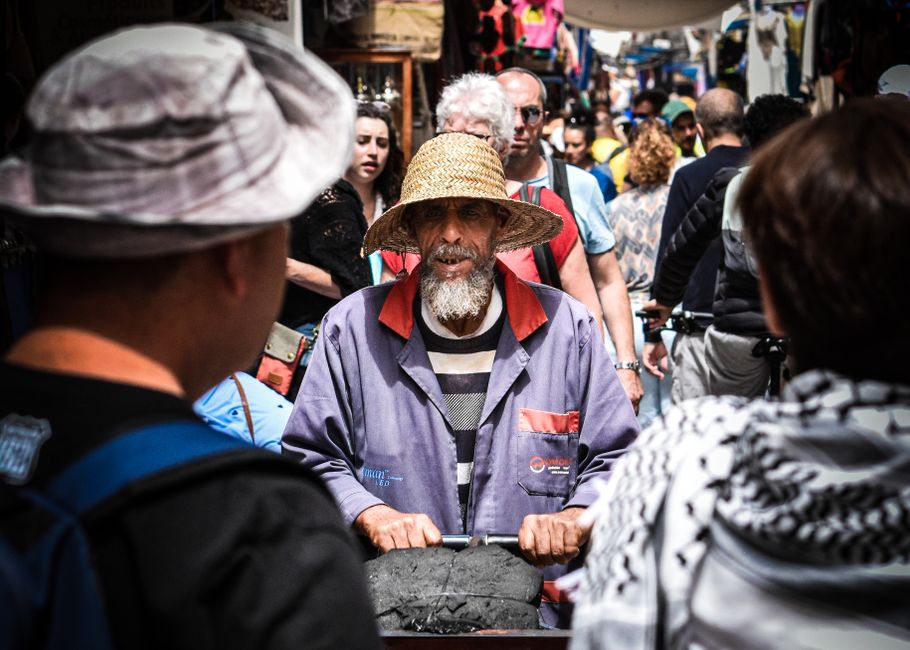 Elderly vendor with straw hat and cart in crowded outdoor market setting.