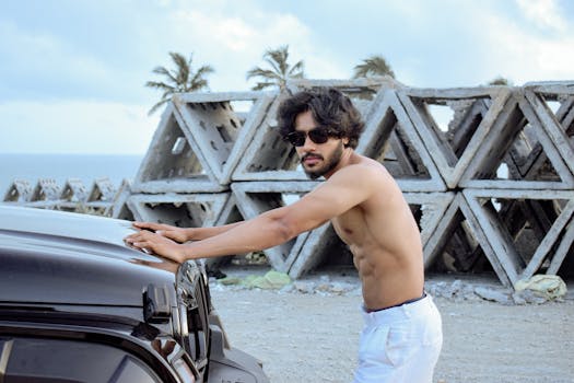 Young man in sunglasses pushing a car by the ocean with concrete structures in the background.