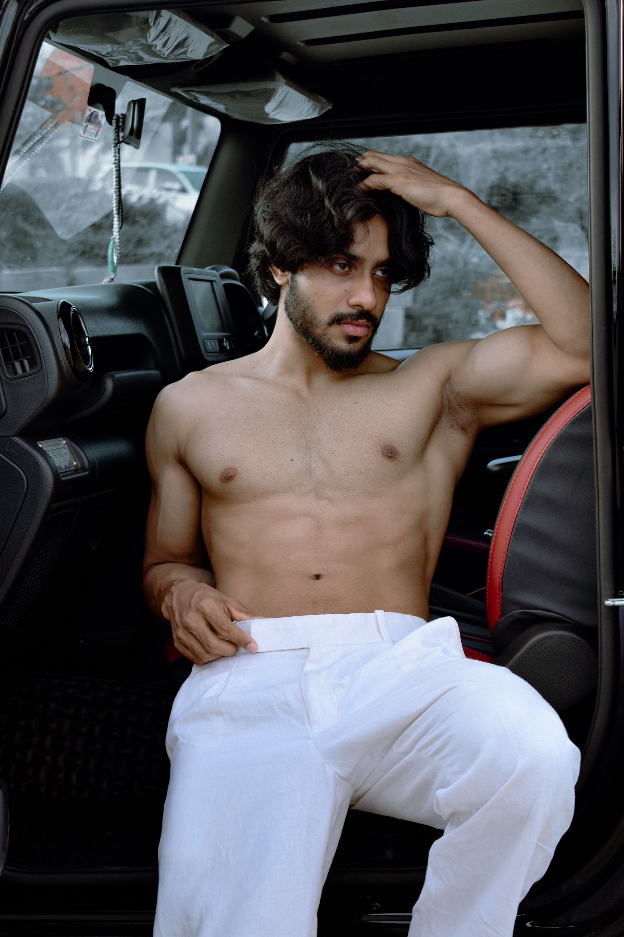 A shirtless man with white pants relaxing in a car seat, portraying a casual and confident look.