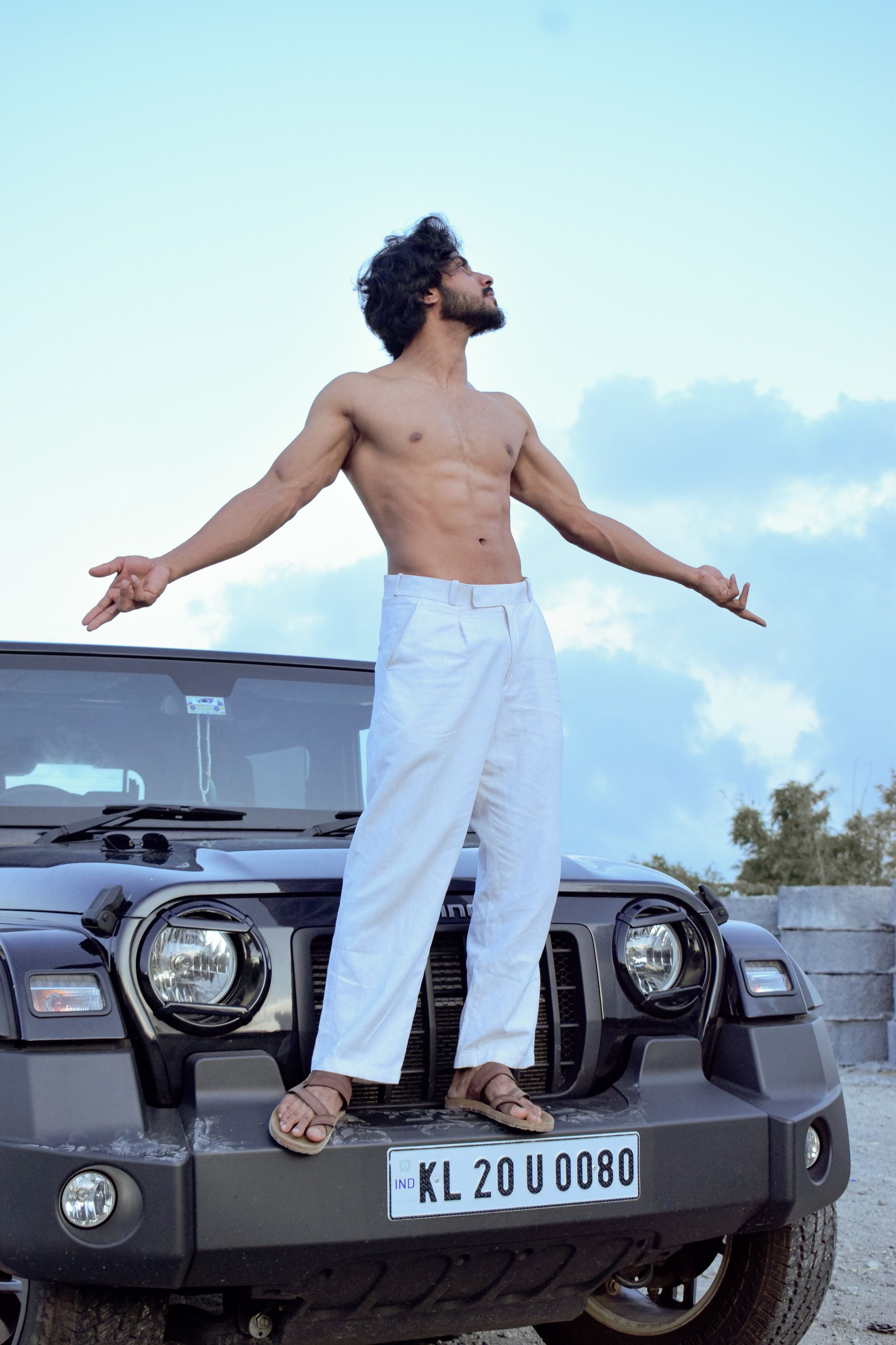 Shirtless man in white pants standing confidently on a black SUV against a blue sky.