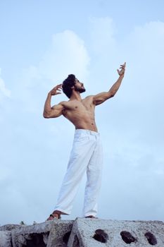 Strong shirtless man poses dramatically atop rocks with a vibrant sky behind.