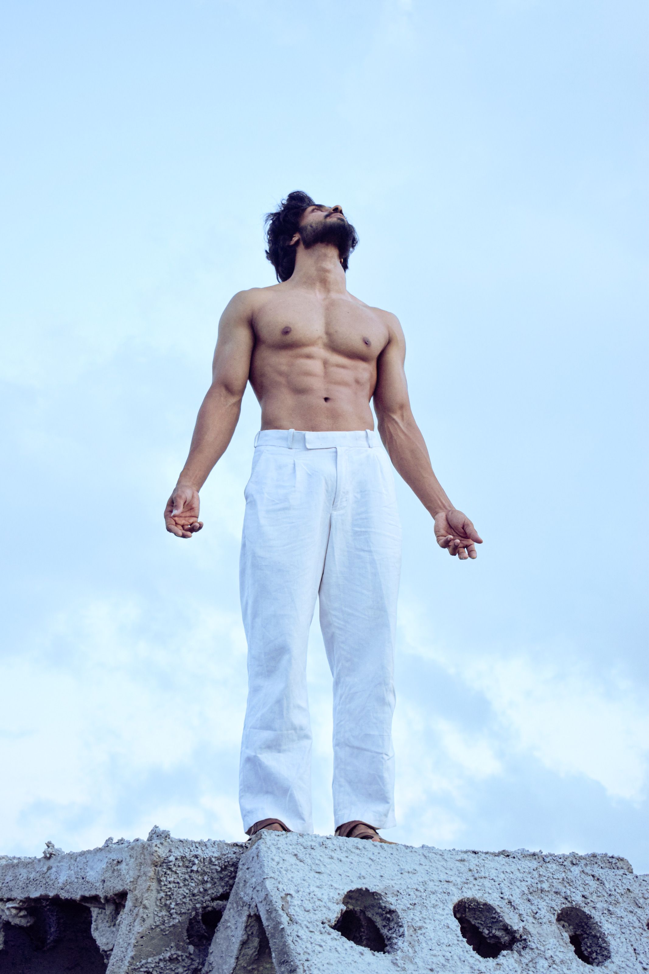 Shirtless man in white pants standing on concrete block looking upwards against clear blue sky.
