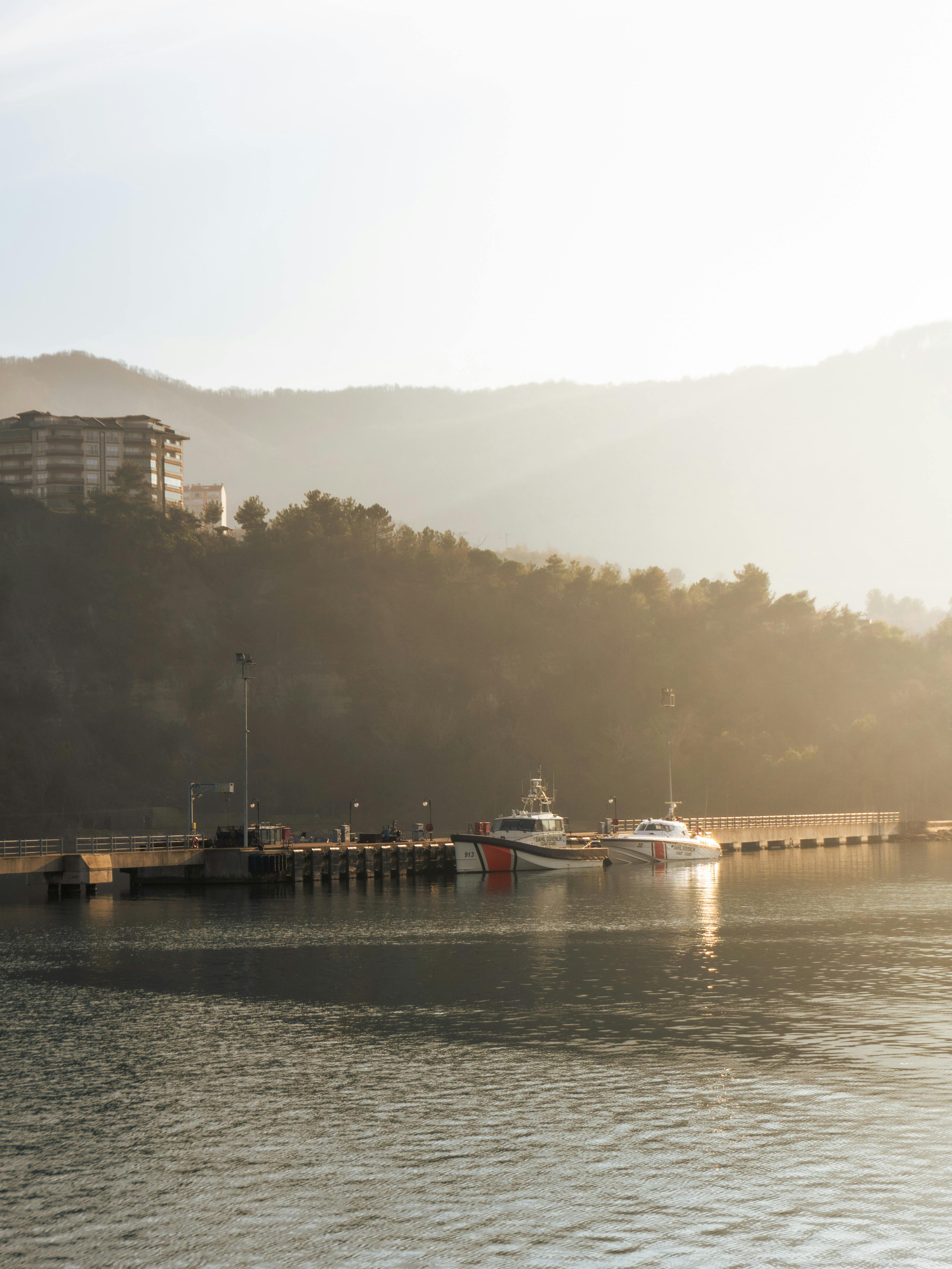 A peaceful lakeside scene with boats docked at a pier during sunrise, surrounded by misty hills.