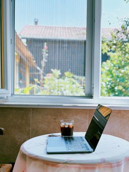 Tranquil home office setup by a window with outdoor view, featuring a laptop and iced coffee on a round table.