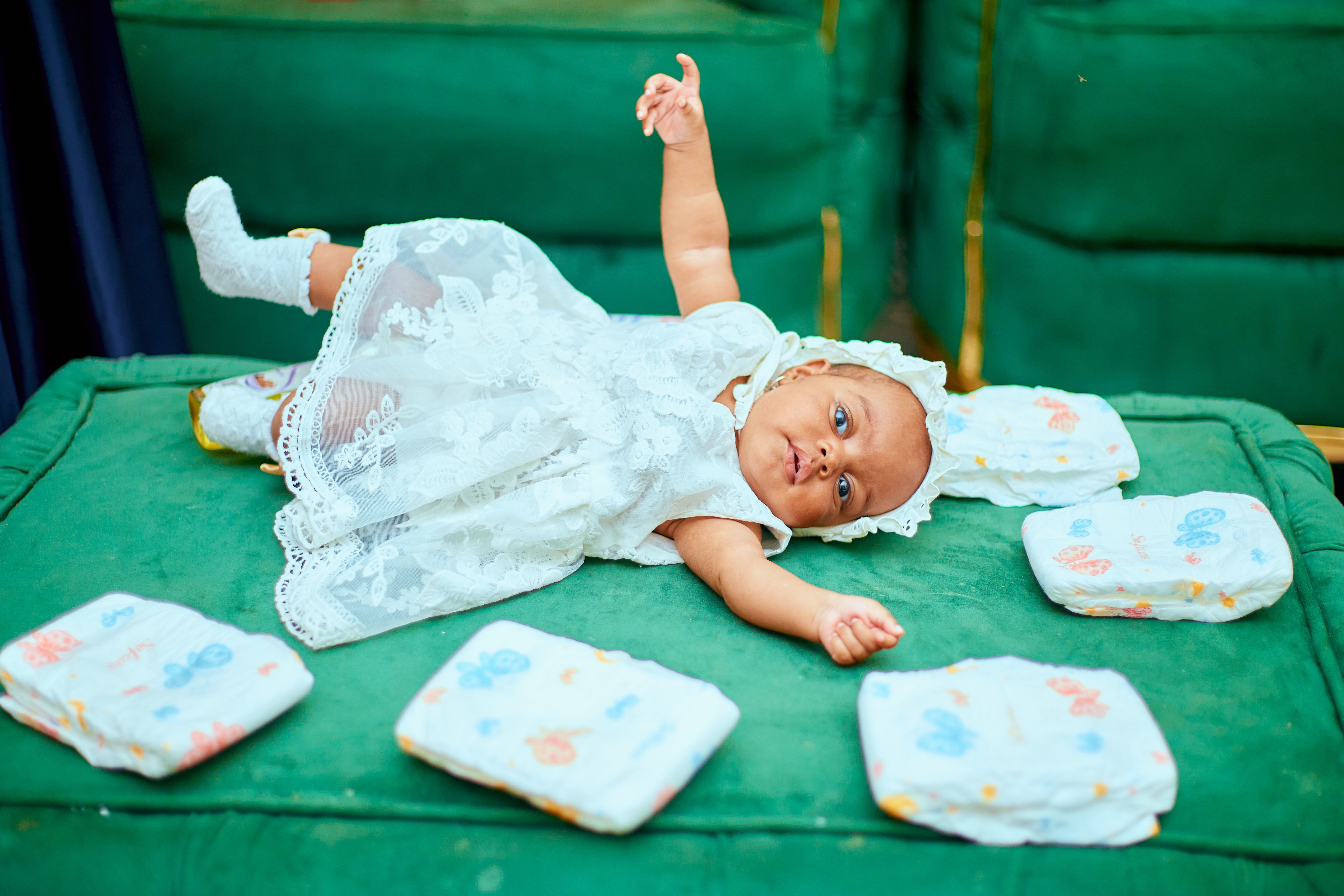 Cute baby in a white dress playing on a green sofa with diapers nearby. Ideal for childcare themes.