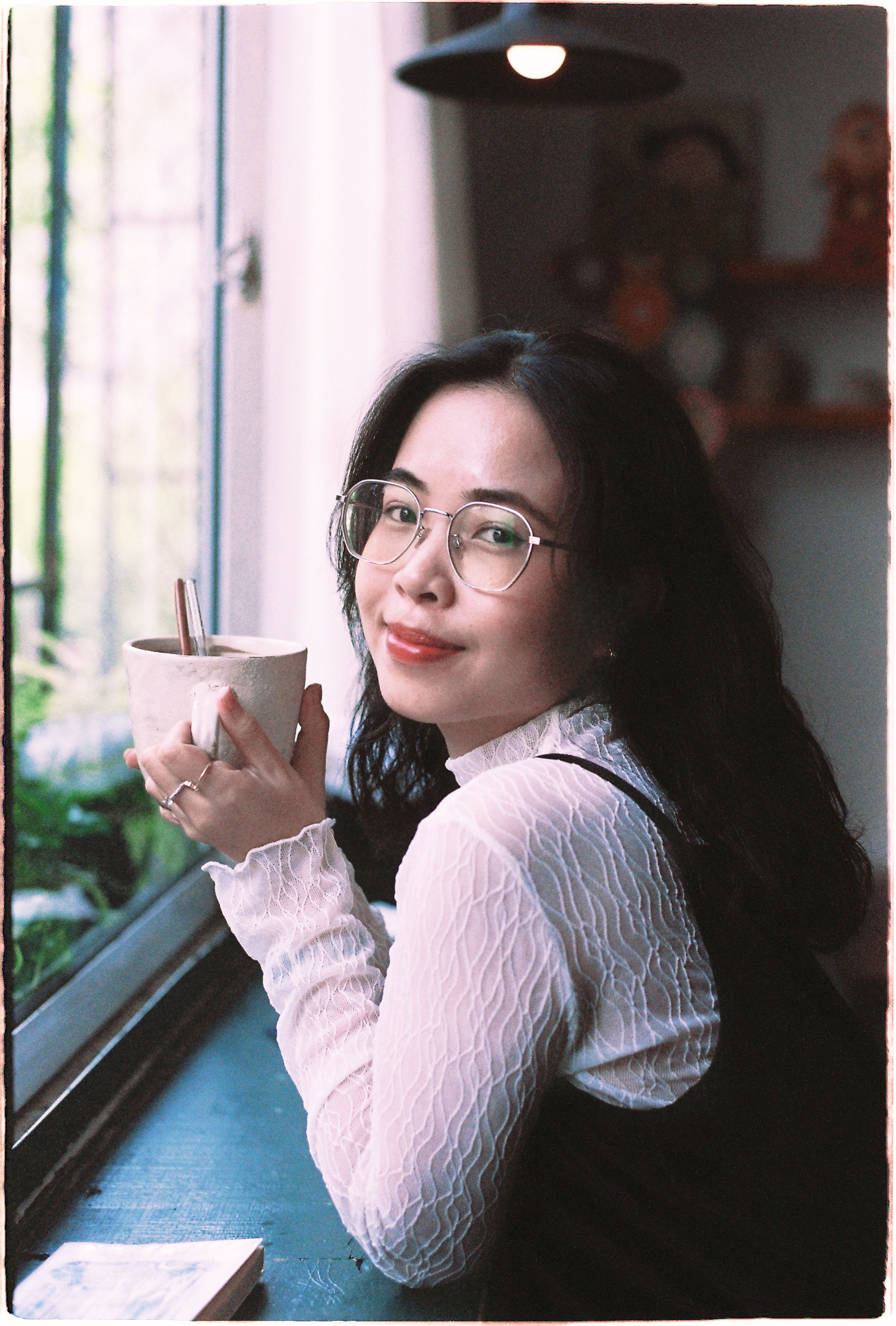 Smiling woman in glasses sipping coffee by a window in a cozy room.