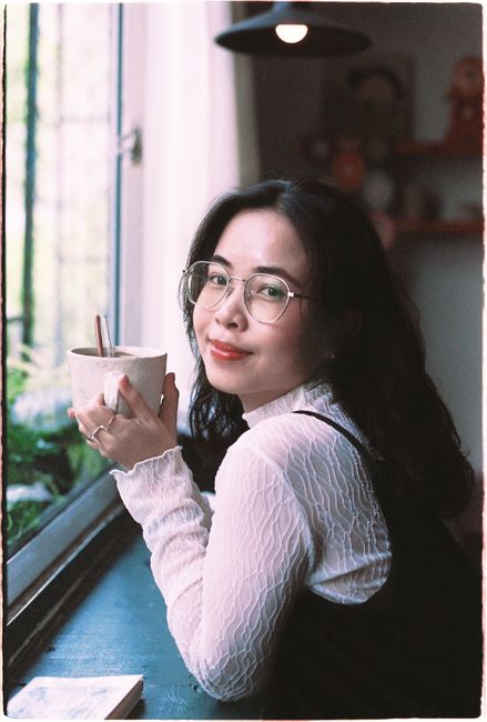 Smiling woman in glasses sipping coffee by a window in a cozy room.