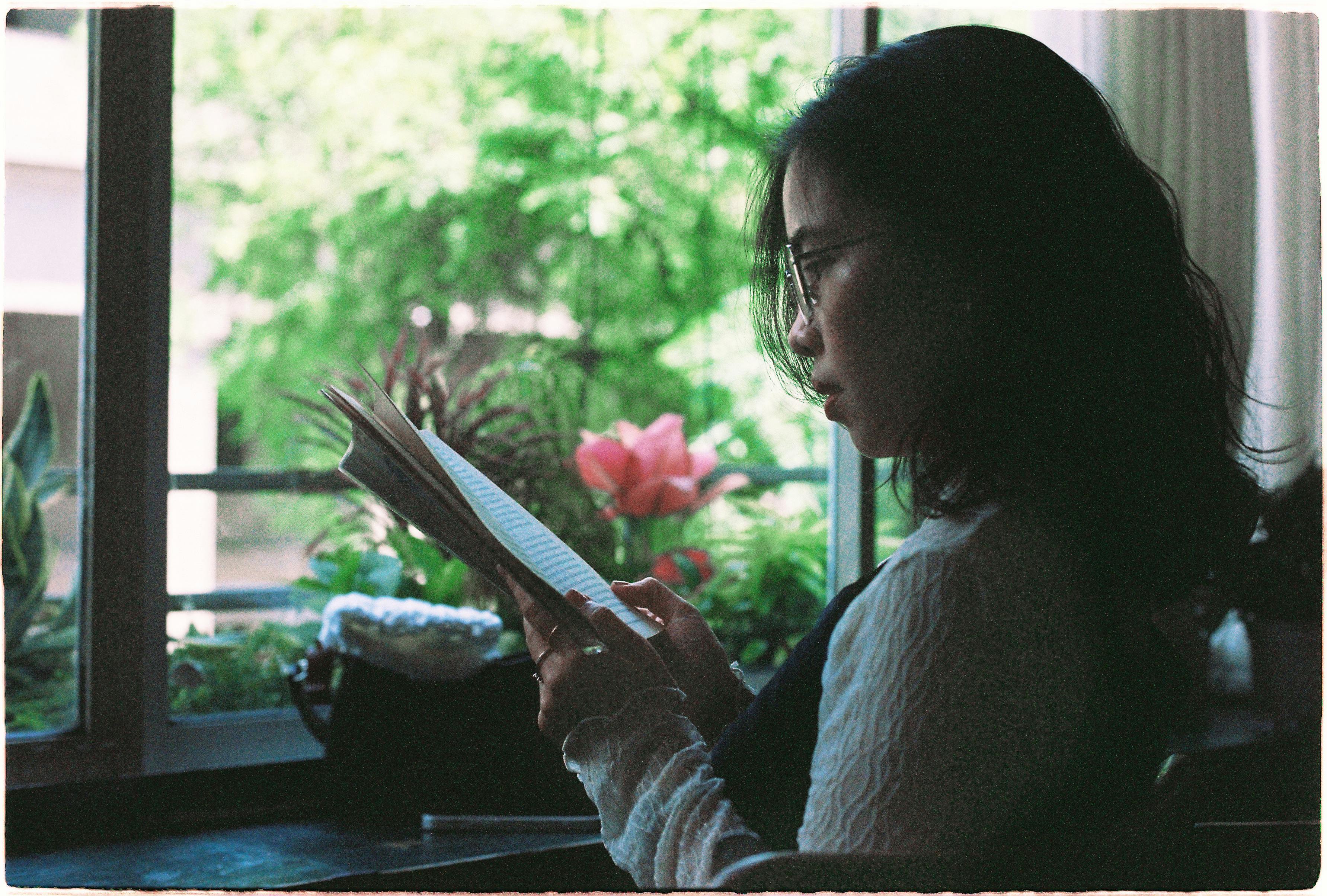 A young woman reads by the window, surrounded by indoor plants in natural daylight.