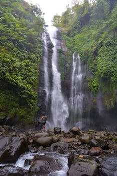 夏日，在高地，一位匿名徒步旅行者站在石头附近，欣赏着从覆盖着绿色植物的岩石悬崖上倾泻而下的风景如画的瀑布