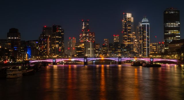 Capture of London's skyline at night with a glowing bridge and reflections on the Thames.