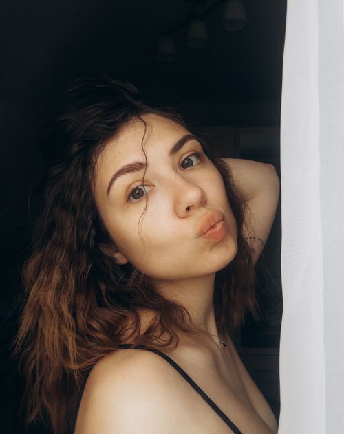Free Portrait of a young woman with a playful expression posing near a sunlit window indoors. Stock Photo