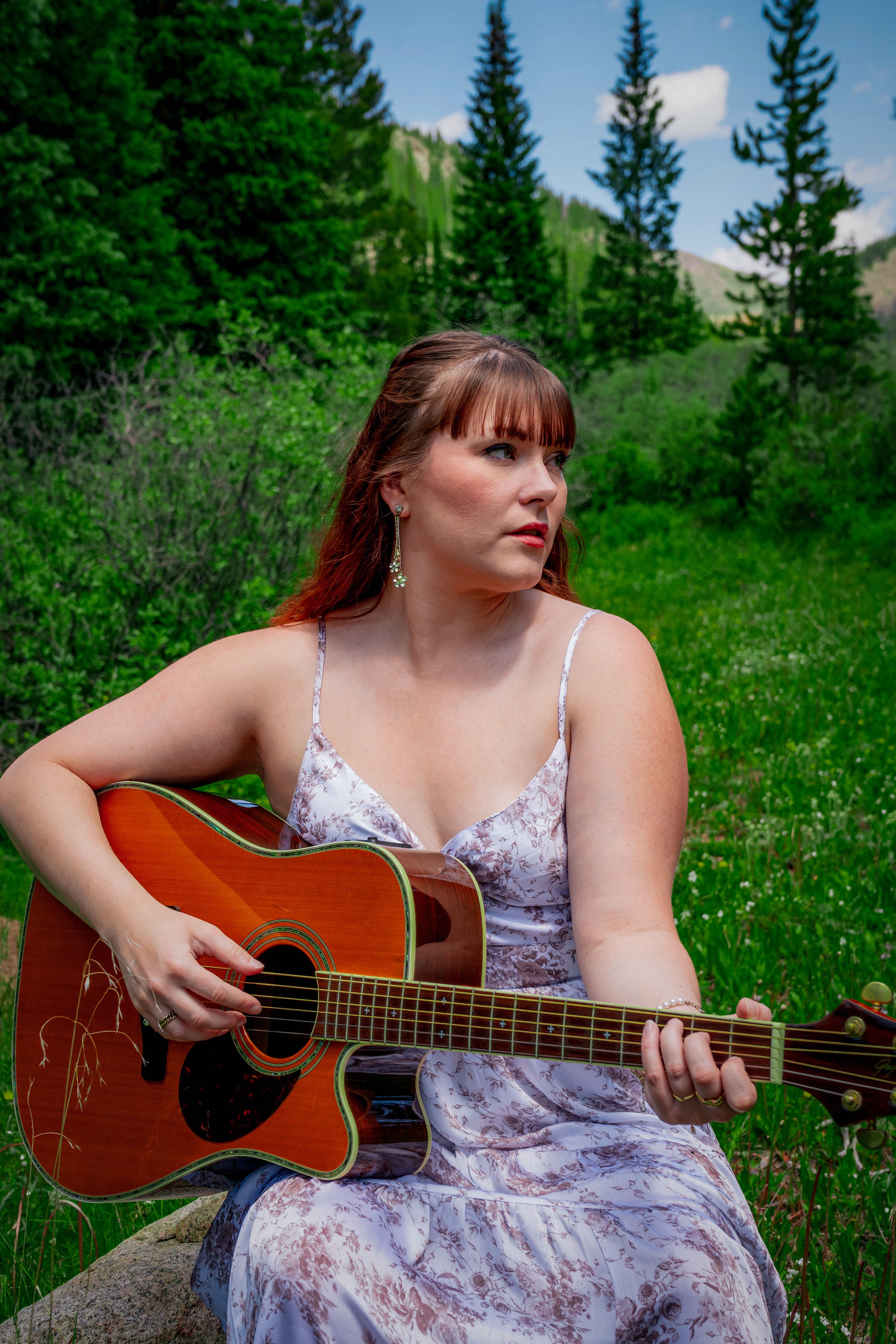 A woman sits outdoors playing an acoustic guitar in a vibrant mountain meadow, exuding a serene and artistic vibe.