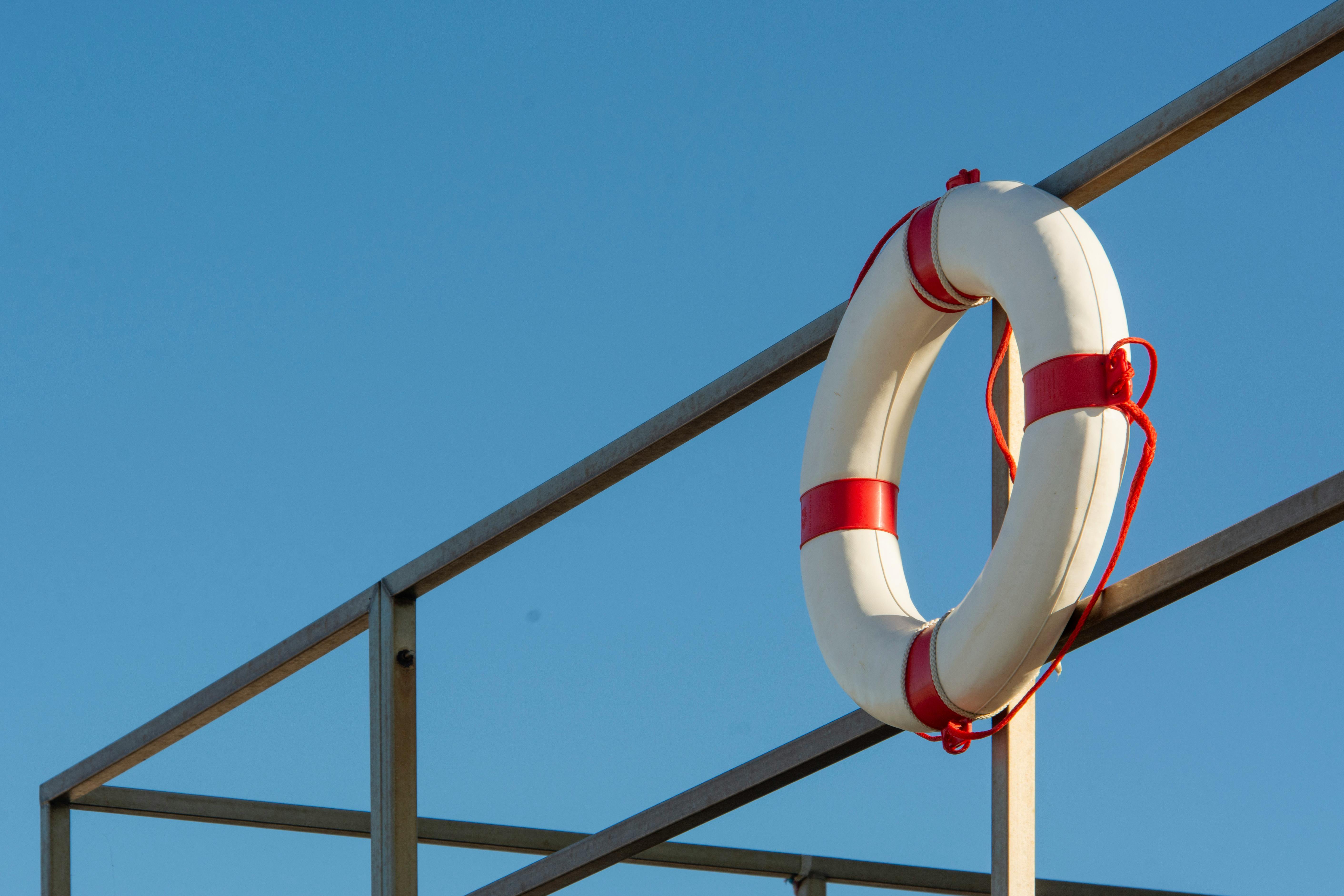A white and red lifebuoy is attached to a metal railing against a clear blue sky, symbolizing safety at sea.