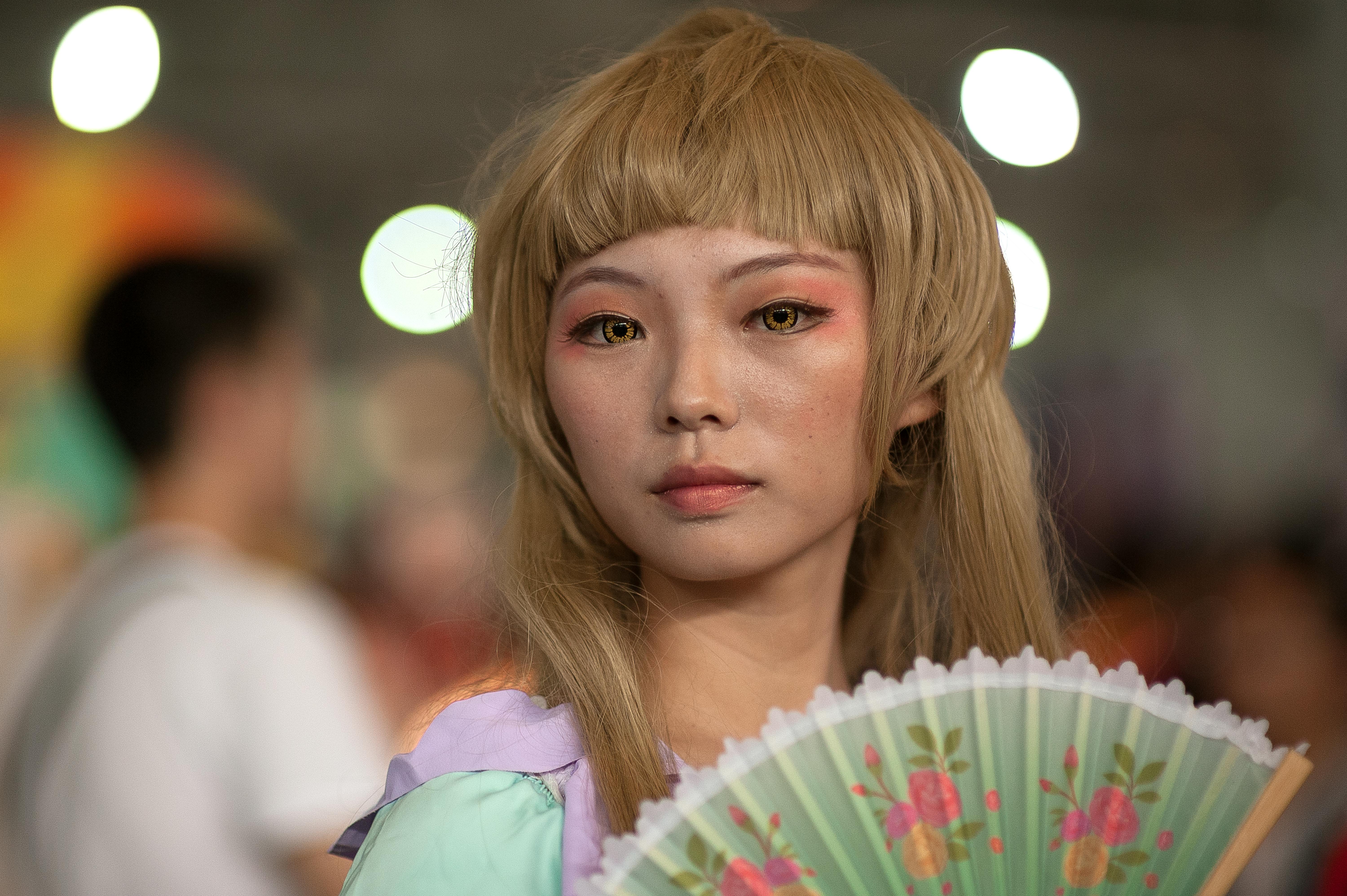 Close-up portrait of a cosplayer holding a fan at an anime convention indoors.