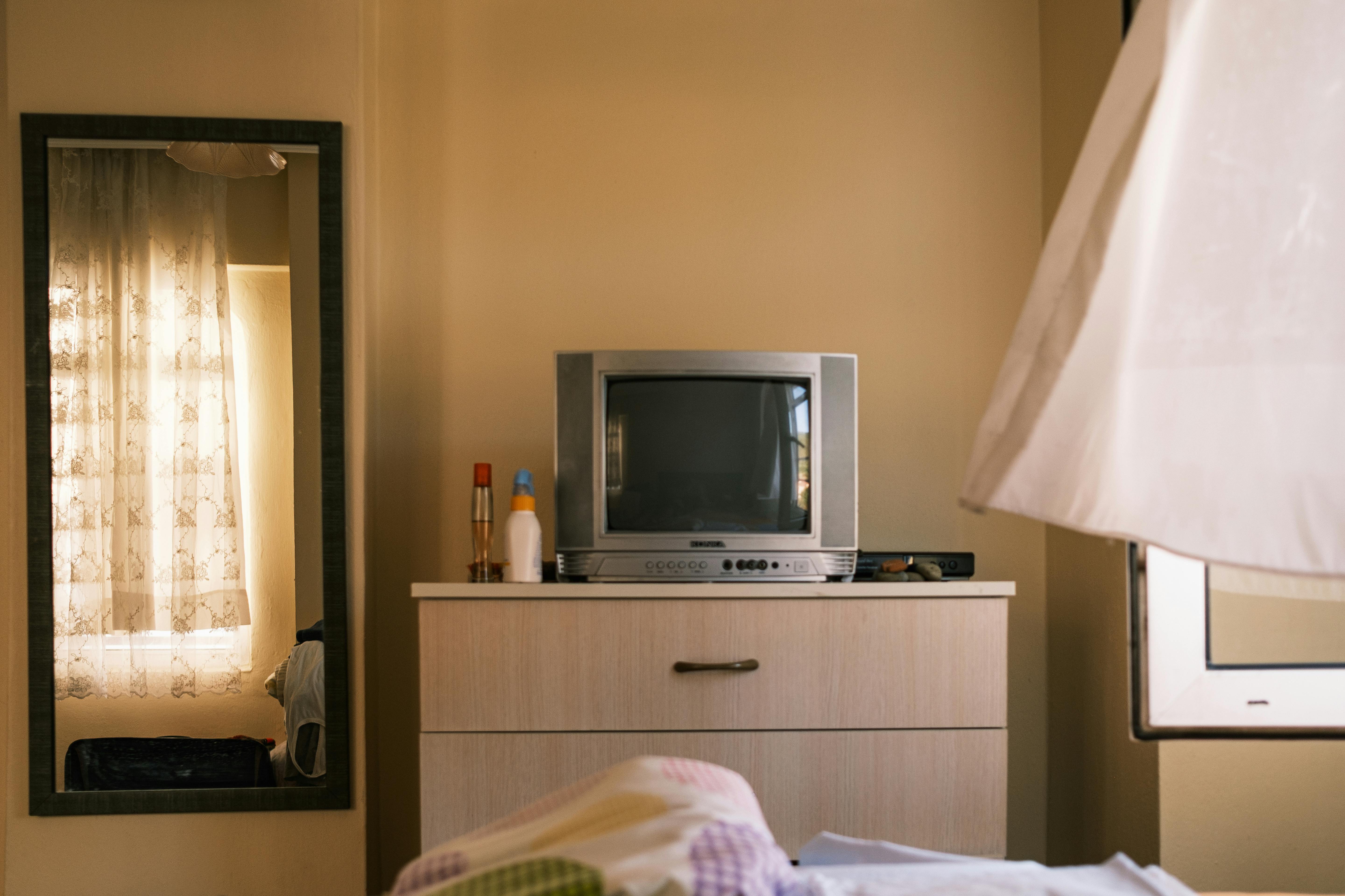 A vintage television on a dresser in a cozy bedroom, featuring a large mirror reflecting sunlight through curtains.