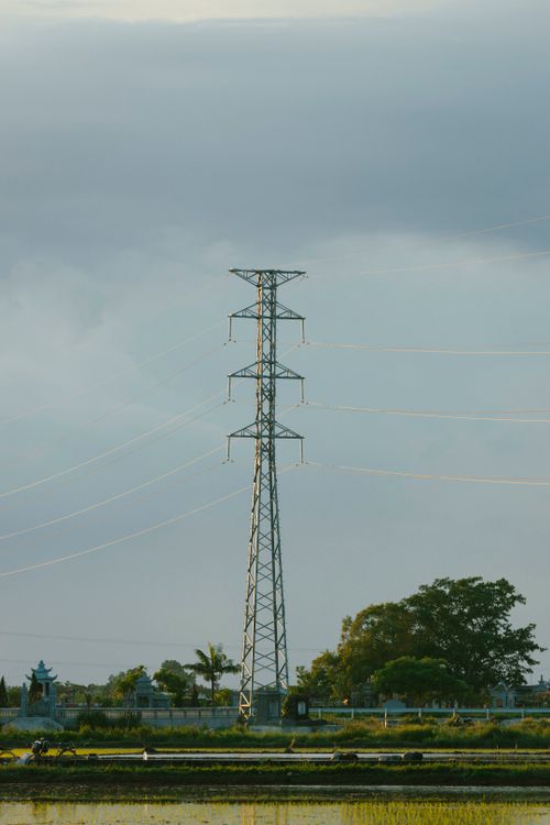 Free A tall transmission tower stands prominently in a rural landscape under a cloudy sky. Stock Photo