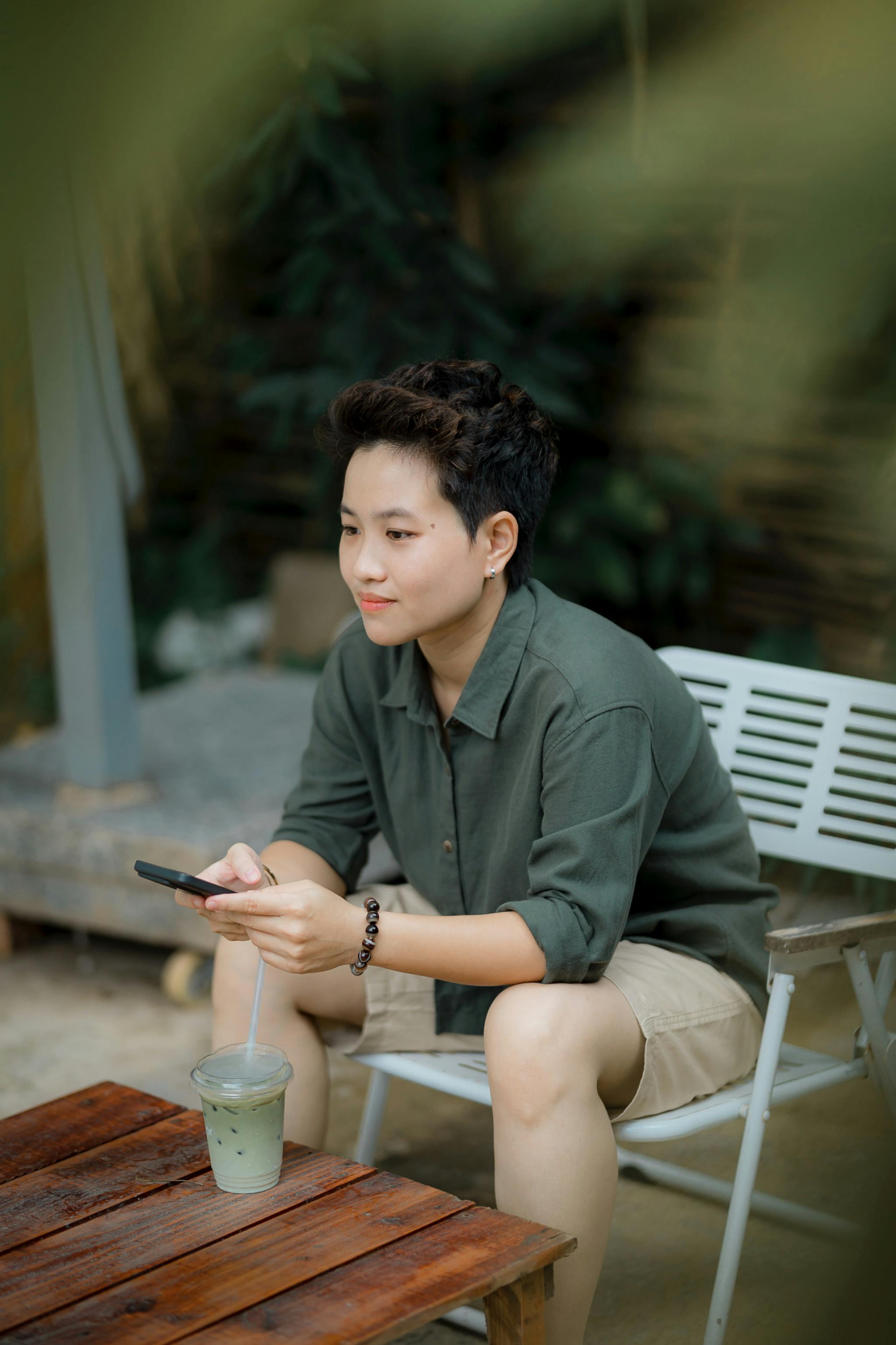 бесплатная Молодой человек сидит на открытом воздухе с телефоном и зеленым напитком на столе, непринужденная обстановка. Стоковое фото